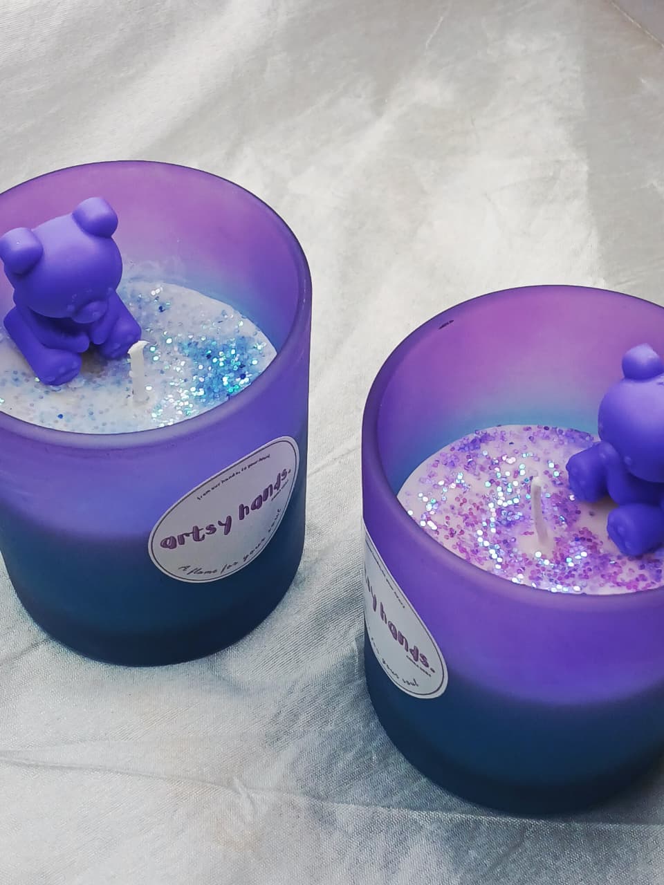 Two purple candles with bear figurines and glitter inside, on a white fabric background.