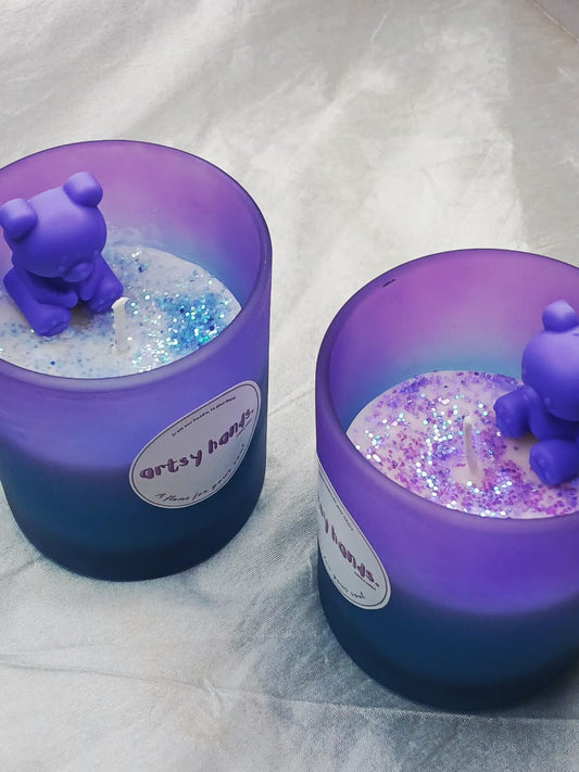 Two purple candles with bear figurines and glitter inside, on a white fabric background.